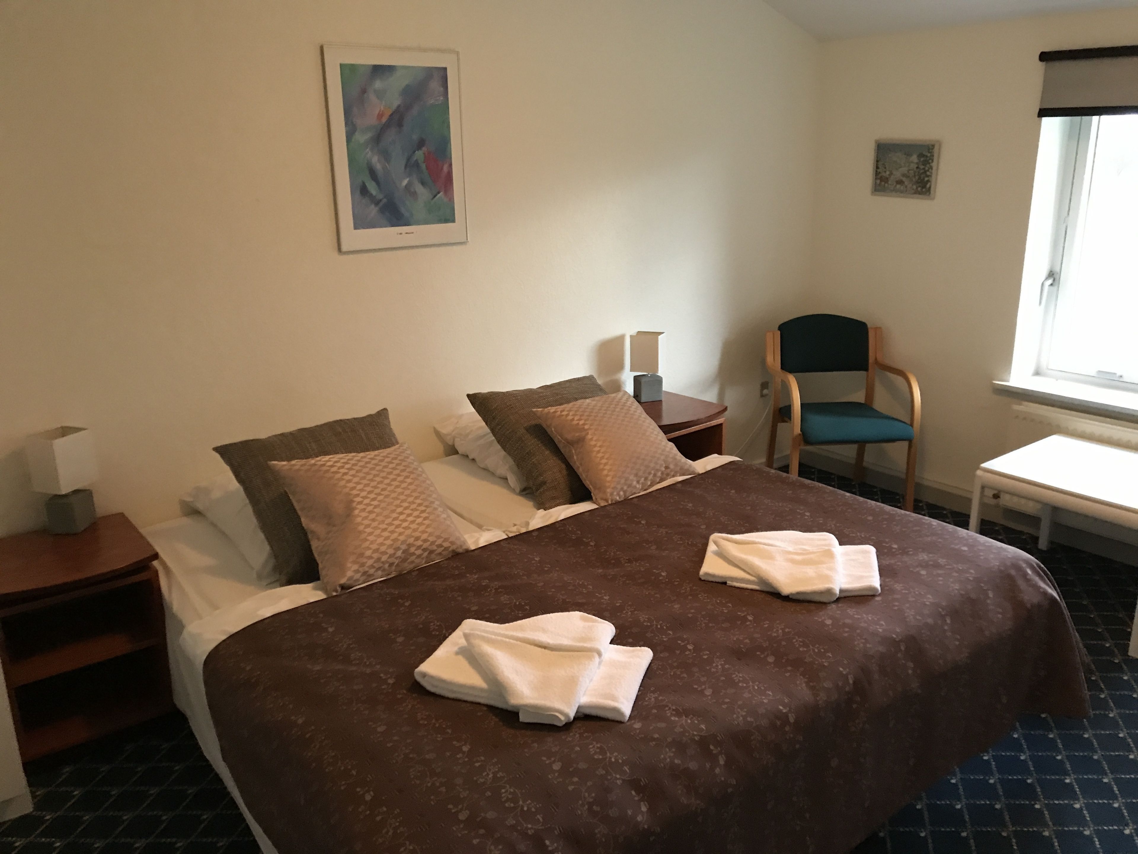 Een hotelkamer met een groot bed, twee kussens, een stoel, een bijzettafeltje en een raam.