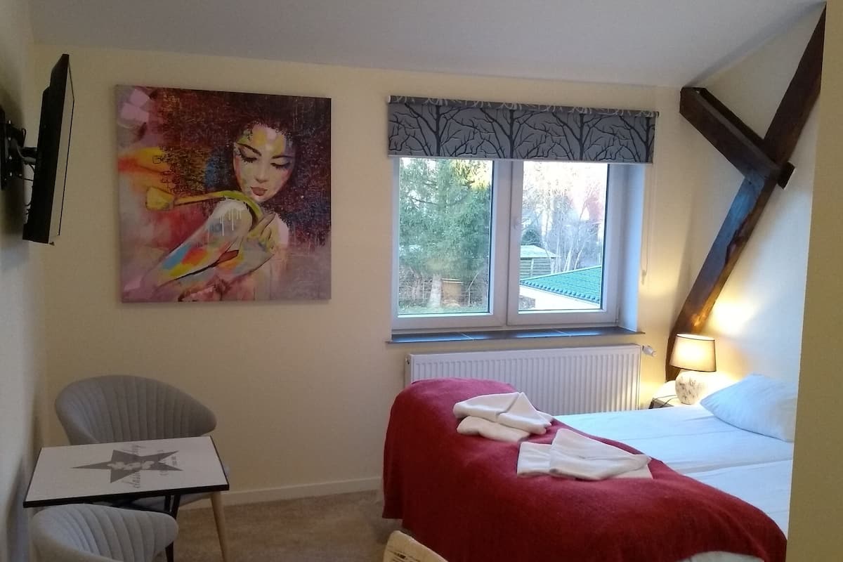 A hotel room with a bed, a chair, a table, a painting, a window, and a radiator.