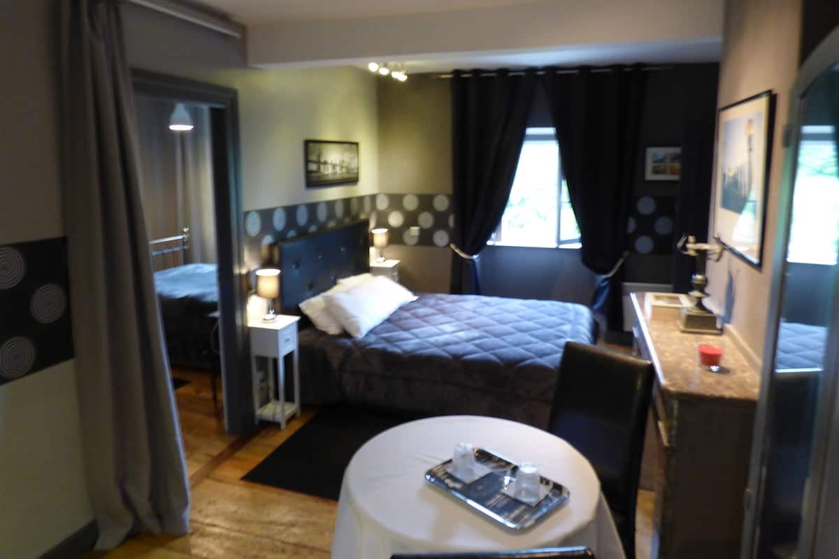 Ein Hotelzimmer mit einem Bett, Nachttischen mit Lampen, einem Schreibtisch mit Stuhl, einem kleinen runden Tisch mit Tablett und einem Fenster mit Vorhängen.