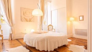 A bedroom with a round bed, wooden flooring, white curtains, a large window, and a wall-mounted lamp.