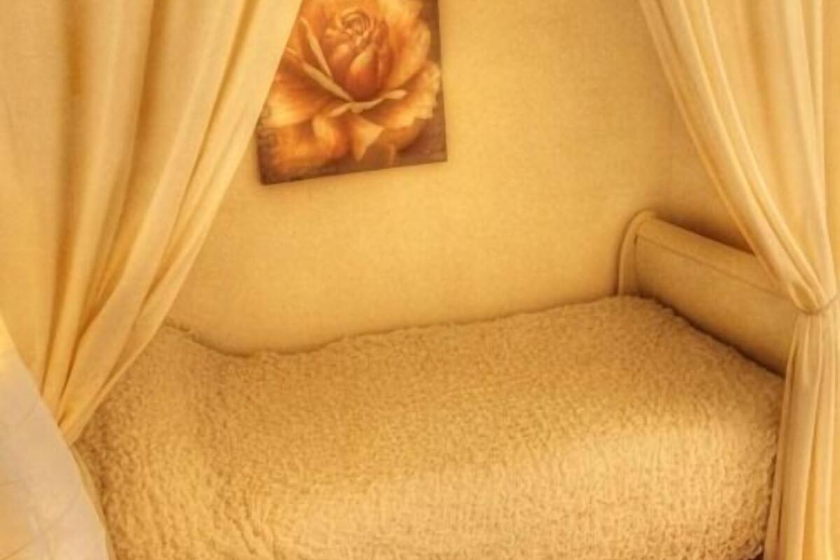 Ein Zimmer mit einem beige gestreiften Bett, einem Rosenbild an der Wand und gelben Vorhängen.