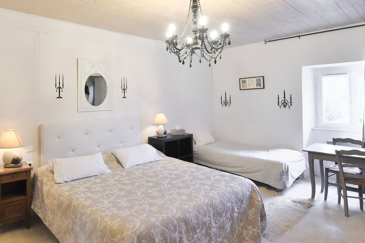 Ein Schlafzimmer mit einem großen Bett, einem kleineren Bett, einem Schreibtisch mit Stuhl und einer Kandelaber.