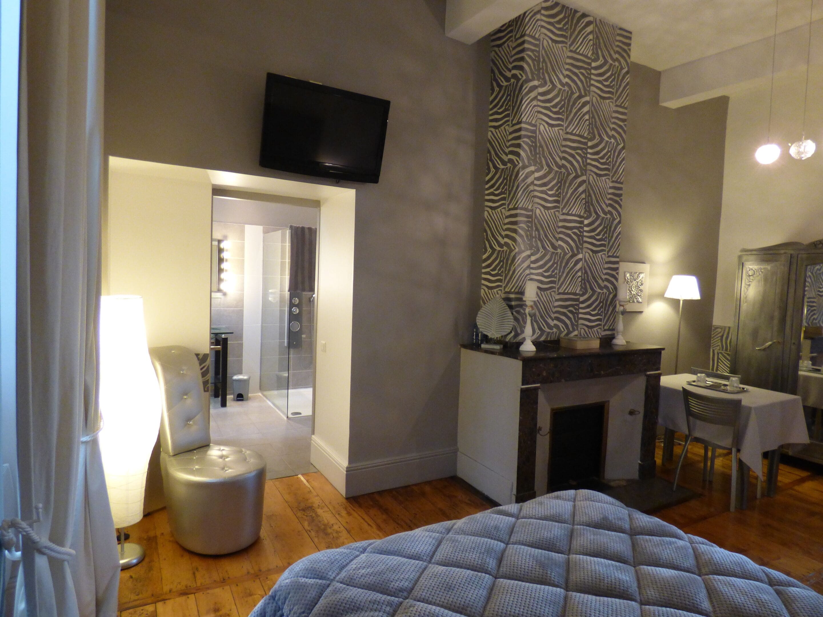 A modern hotel room with a large bed, a flat-screen TV mounted on the wall, a zebra-patterned accent wall, a small table with a lamp, and a fireplace.
