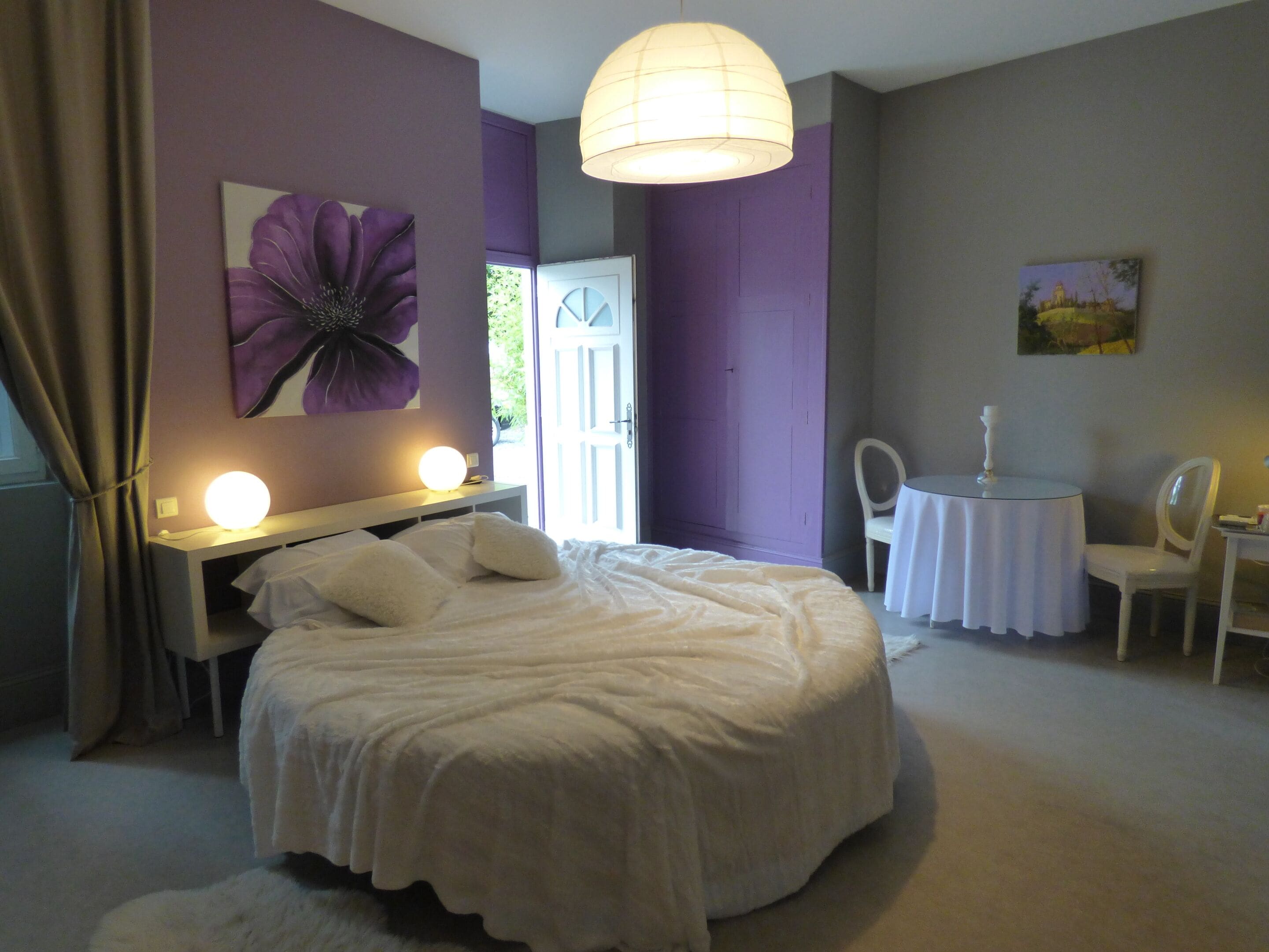 A bedroom with a large bed, a bedside table with a lamp, a chair, a small round table with a lamp, and a door leading to another room.