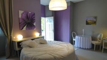 A bedroom with a large bed, a bedside table with a lamp, a chair, a small round table with a lamp, and a door leading to another room.