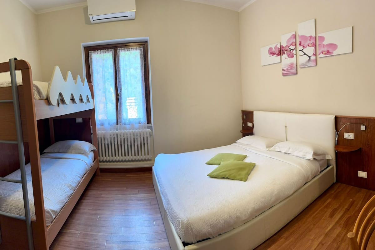 Quarto com beliches, radiador, janela com cortinas, mesa de cabeceira, cadeira e ar-condicionado fixado na parede.