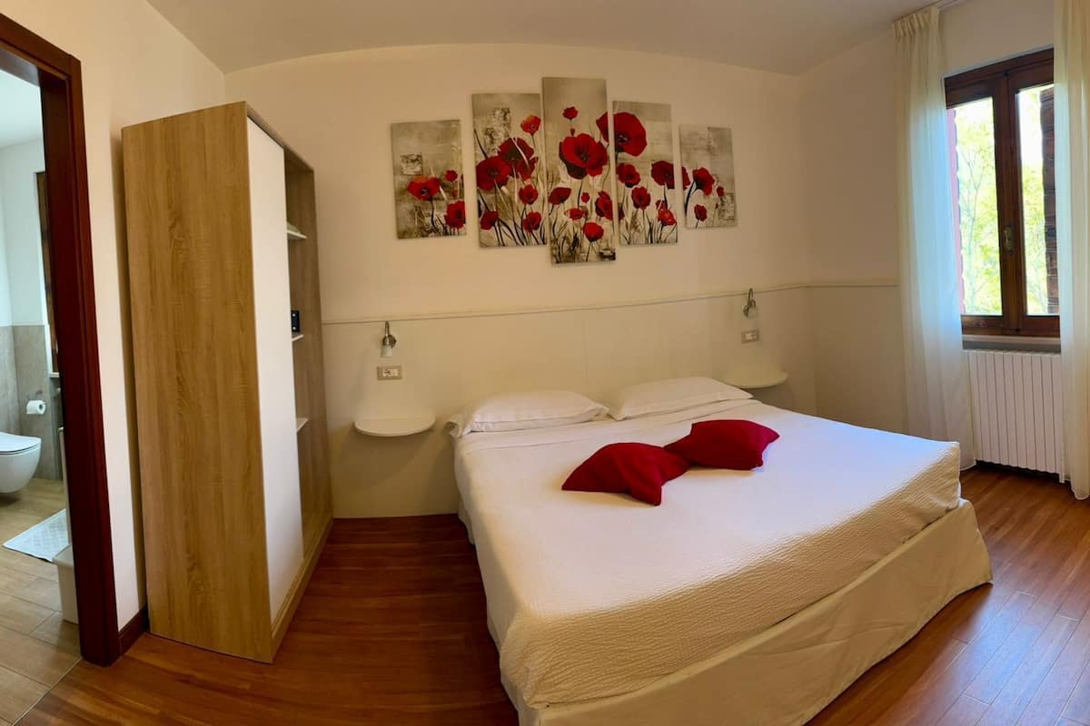Um quarto com piso de madeira, cama com roupa de cama branca e almofadas vermelhas, banheiro com vaso sanitário e pia, e uma janela com cortinas.