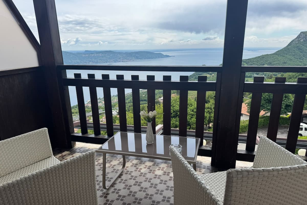 Uma varanda com cadeiras de vime e uma mesa, com vista para a paisagem costeira e montanhosa.