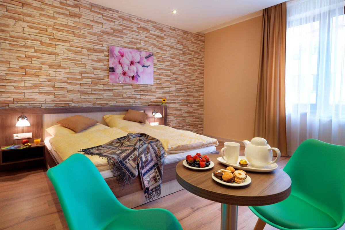 A hotel room with a bed, two green chairs, a table with breakfast items, a painting on the wall, and a window with curtains.