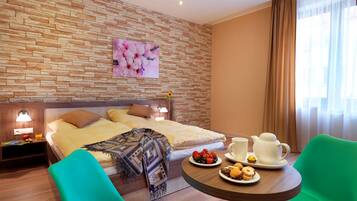 A hotel room with a bed, two green chairs, a table with breakfast items, a painting on the wall, and a window with curtains.