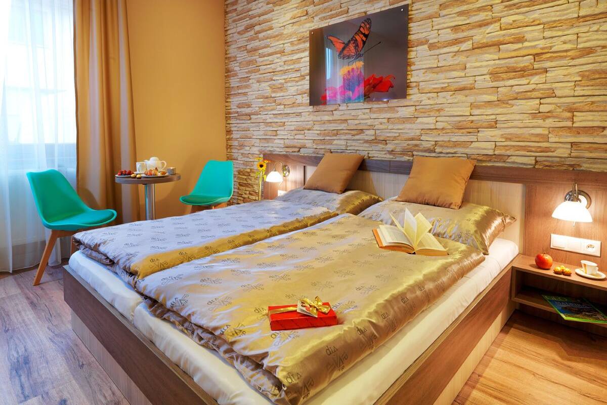 A hotel room with a large bed, two chairs, a small table with a drink, and a wall-mounted lamp.