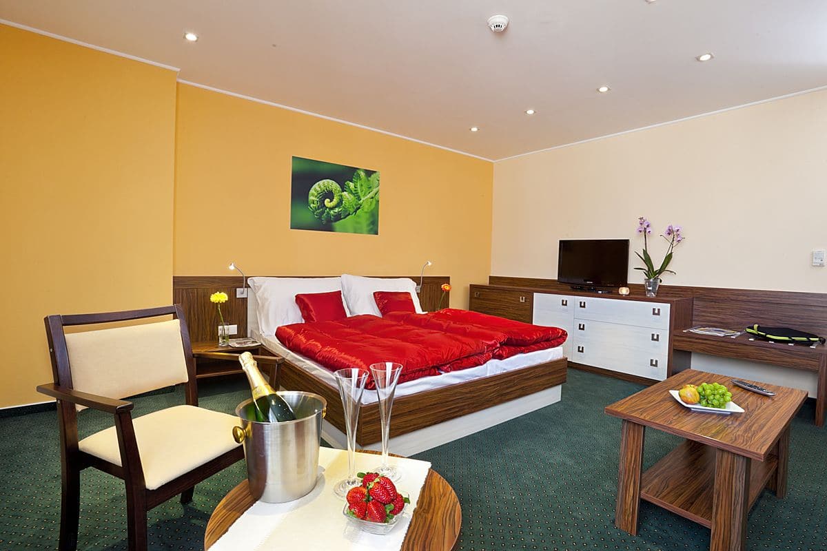 A hotel room with a bed, a desk, a chair, a small table with fruit, a bottle of champagne, and a vase with flowers.