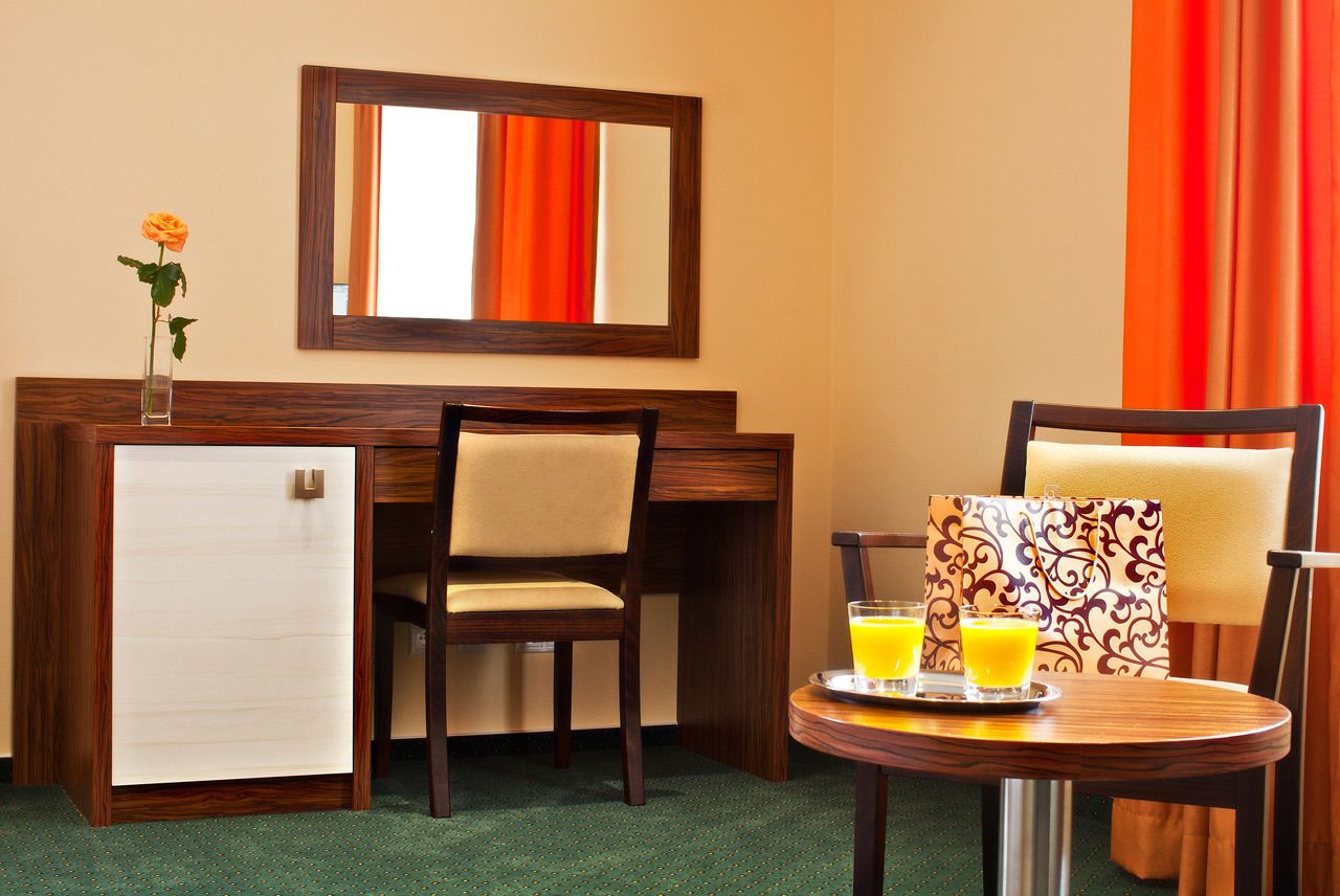 A hotel room with a wooden desk, a small refrigerator, a chair, a table with a tray of orange juice, and a mirror on the wall.