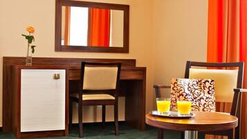 Ein Hotelzimmer mit Schreibtisch aus Holz, einem kleinen Kühlschrank, einem Sessel, einem Tisch mit Orangensaft und einem Spiegel an der Wand.