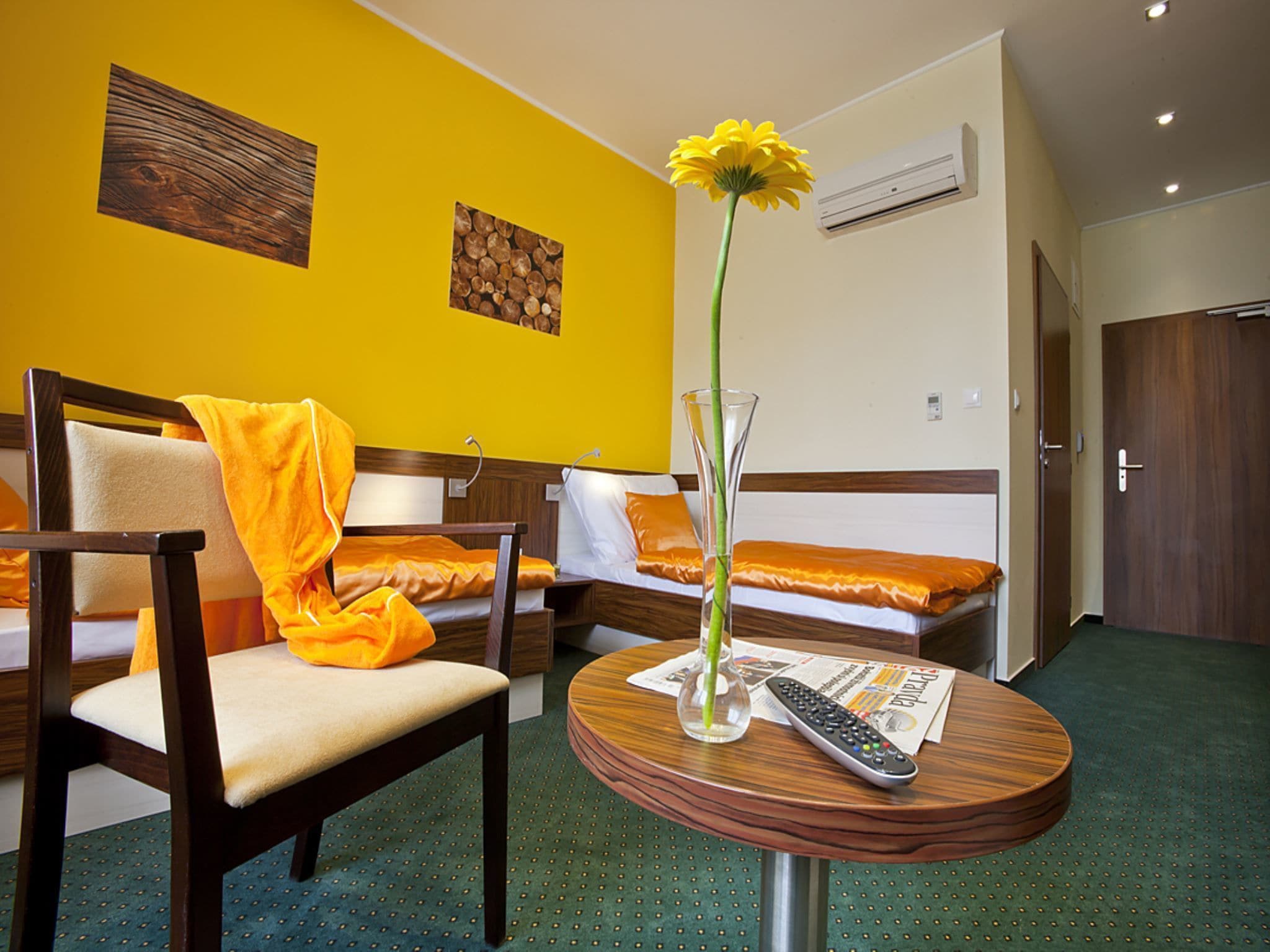 Habitación de hotel con una silla de madera, una mesa redonda con un florero y control remoto, y dos camas con ropa de cama naranja.