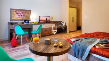 Une chambre d’hôtel moderne avec un plancher en bois, une table ronde avec des collations et un verre de vin, un lit avec une couverture rouge et un bureau avec un ordinateur et une télévision.