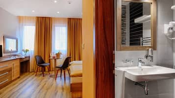 A hotel room with a wooden floor, a bed, a desk with a chair, a small table with a vase of flowers, and a bathroom with a sink and mirror.