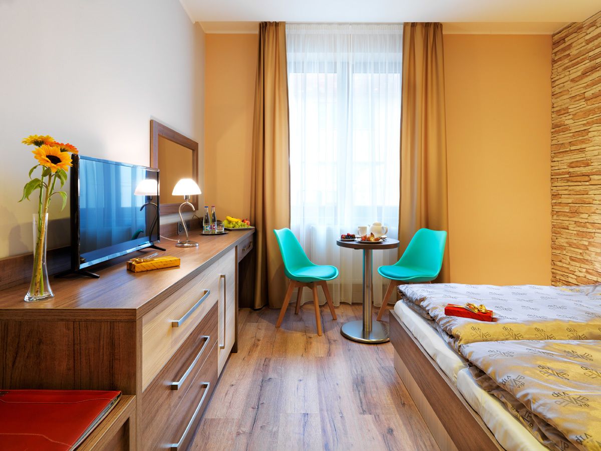 Habitación de hotel con una cama, un escritorio con televisión, dos sillas, una mesita y una ventana con cortinas.