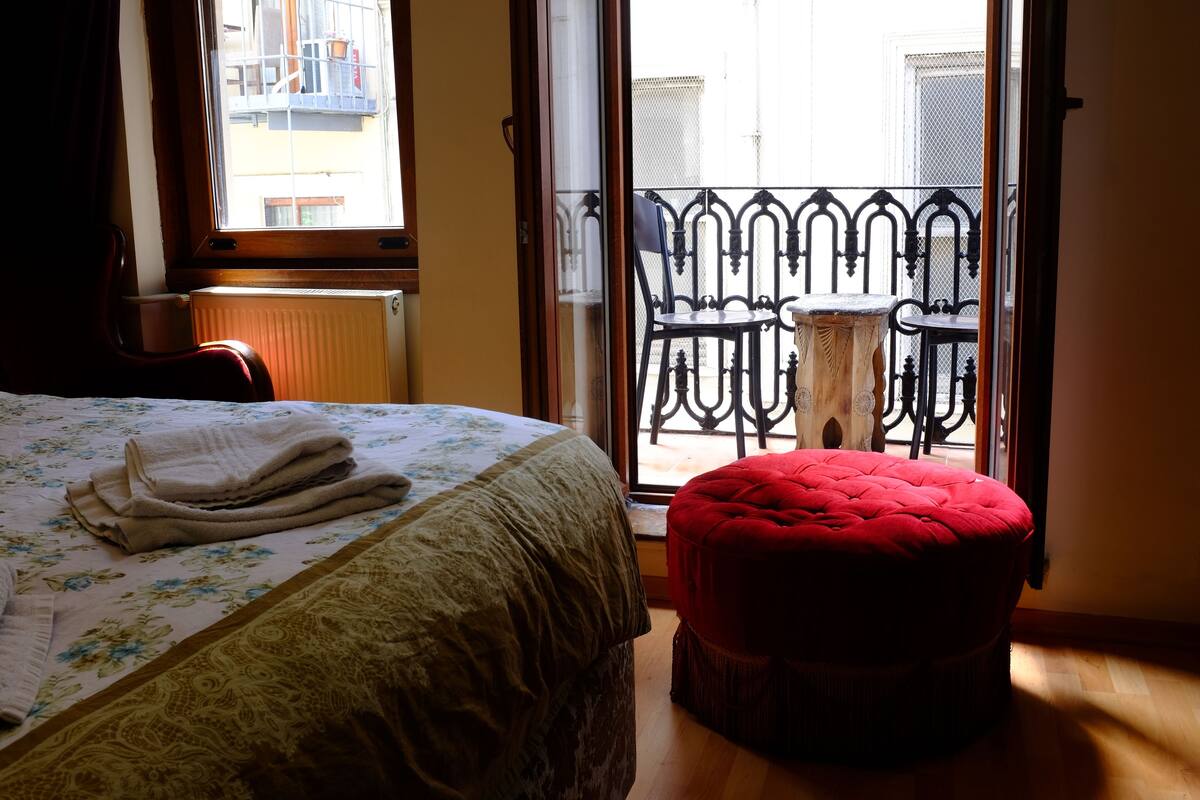 A bedroom with a bed, a red ottoman, a window, and a balcony with a metal railing.