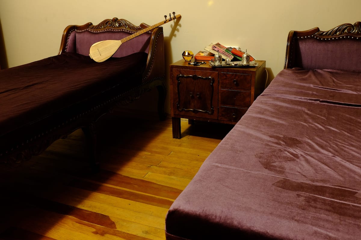 A room with two beds, one with a wooden headboard and the other with a purple headboard. There is a small wooden nightstand with a lamp and a few items on it.
