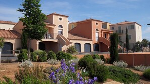 Un edificio residenziale con diversi balconi, un giardino con fiori viola e un cielo azzurro.