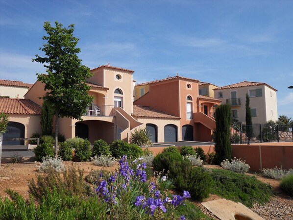 A residential building with multiple balconies, a garden with purple flowers, and a clear blue sky. - Residence Village d'Oc (Béziers)