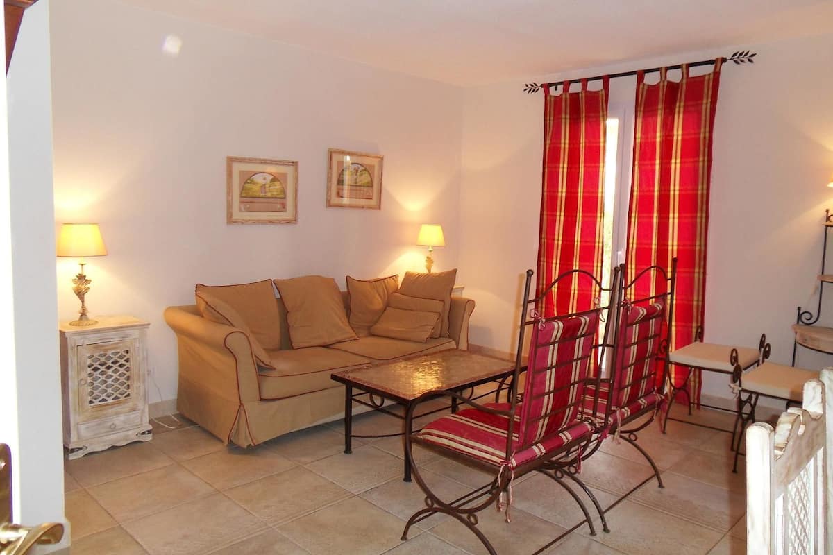 A cozy living room with a sofa, a wooden table, and a metal chair. There are two lamps on side tables and a red curtain.