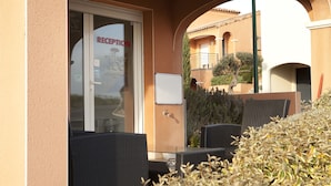 Area esterna con posti a sedere, sedie in vimini, un tavolo, una segnaletica della reception e un edificio con archi e un balcone.