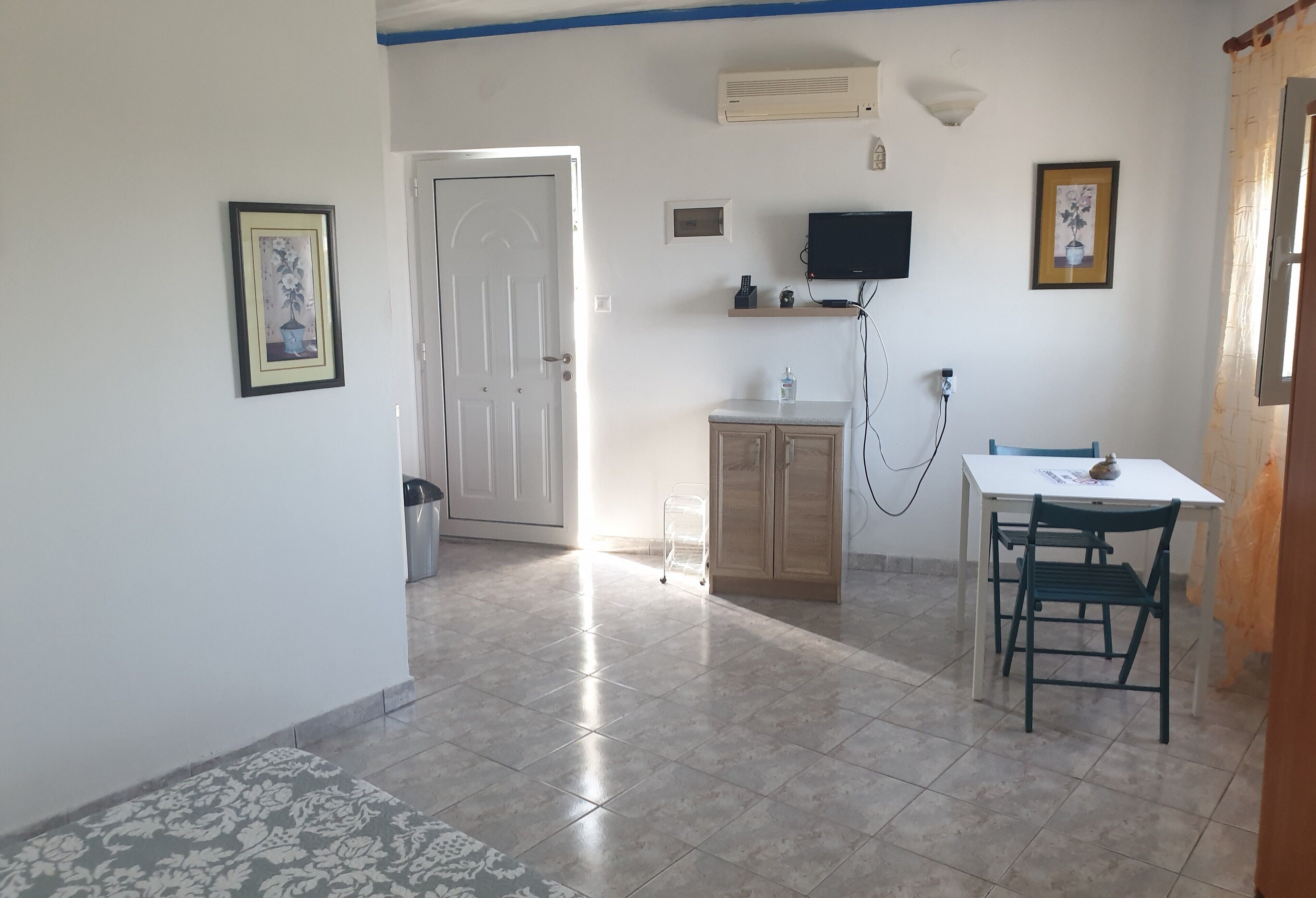 A room with a tiled floor, a white door, a small cabinet, a TV mounted on the wall, and a table with chairs.