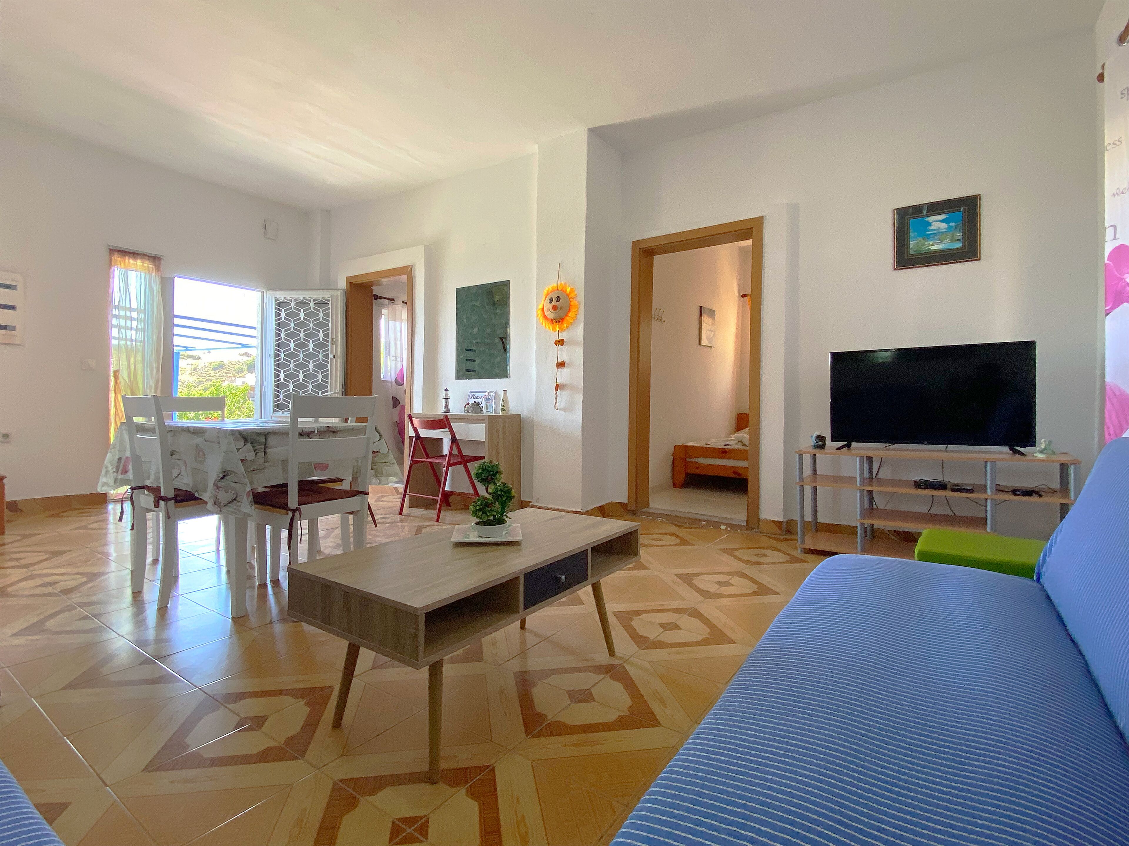 Una sala de estar con un sofá, un televisor, una mesa de comedor con sillas y una planta pequeña sobre una mesa de centro.
