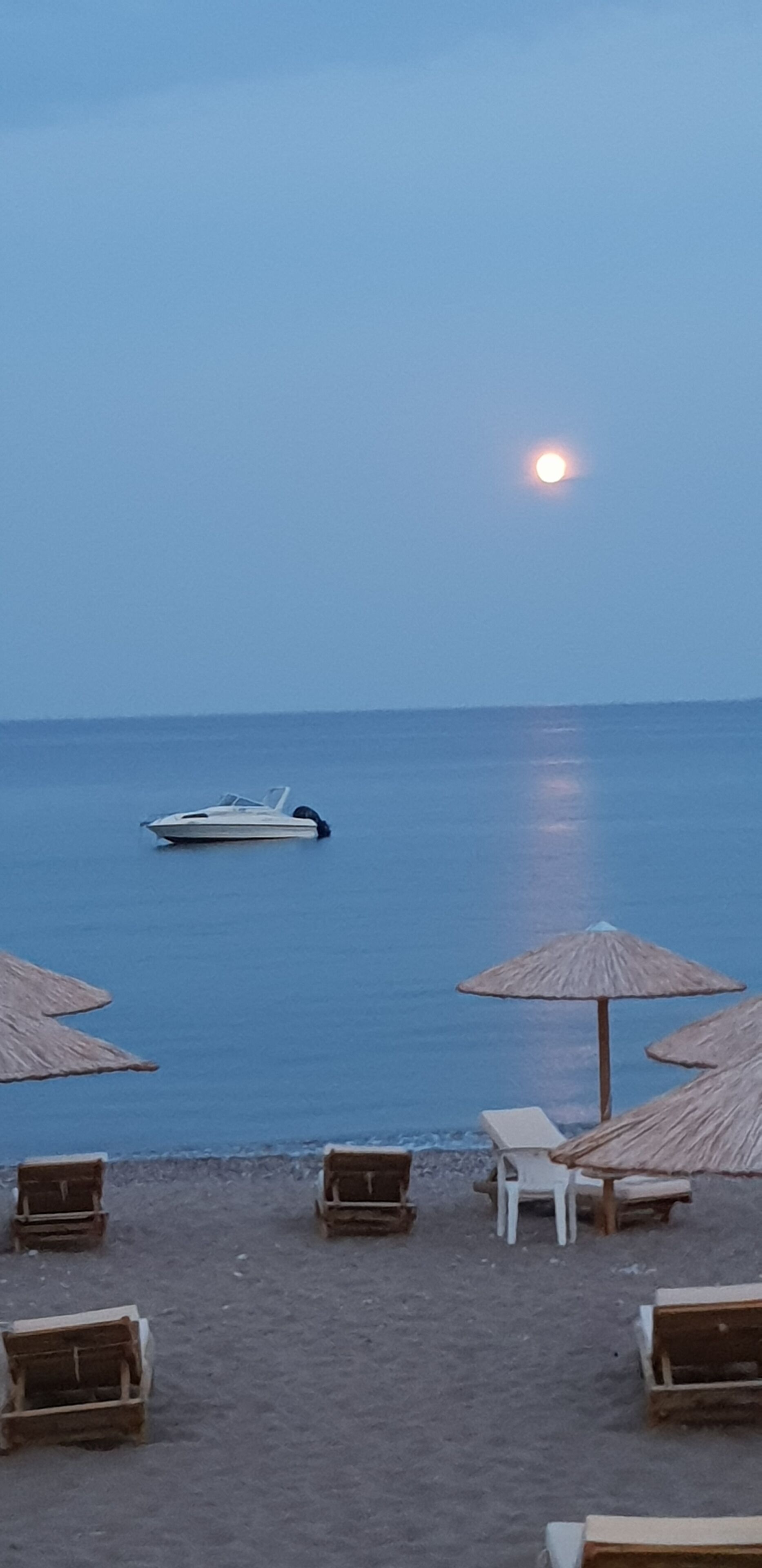 Una spiaggia con sedie a sdraio e ombrelloni, una barca sull'acqua e un tramonto sullo sfondo.