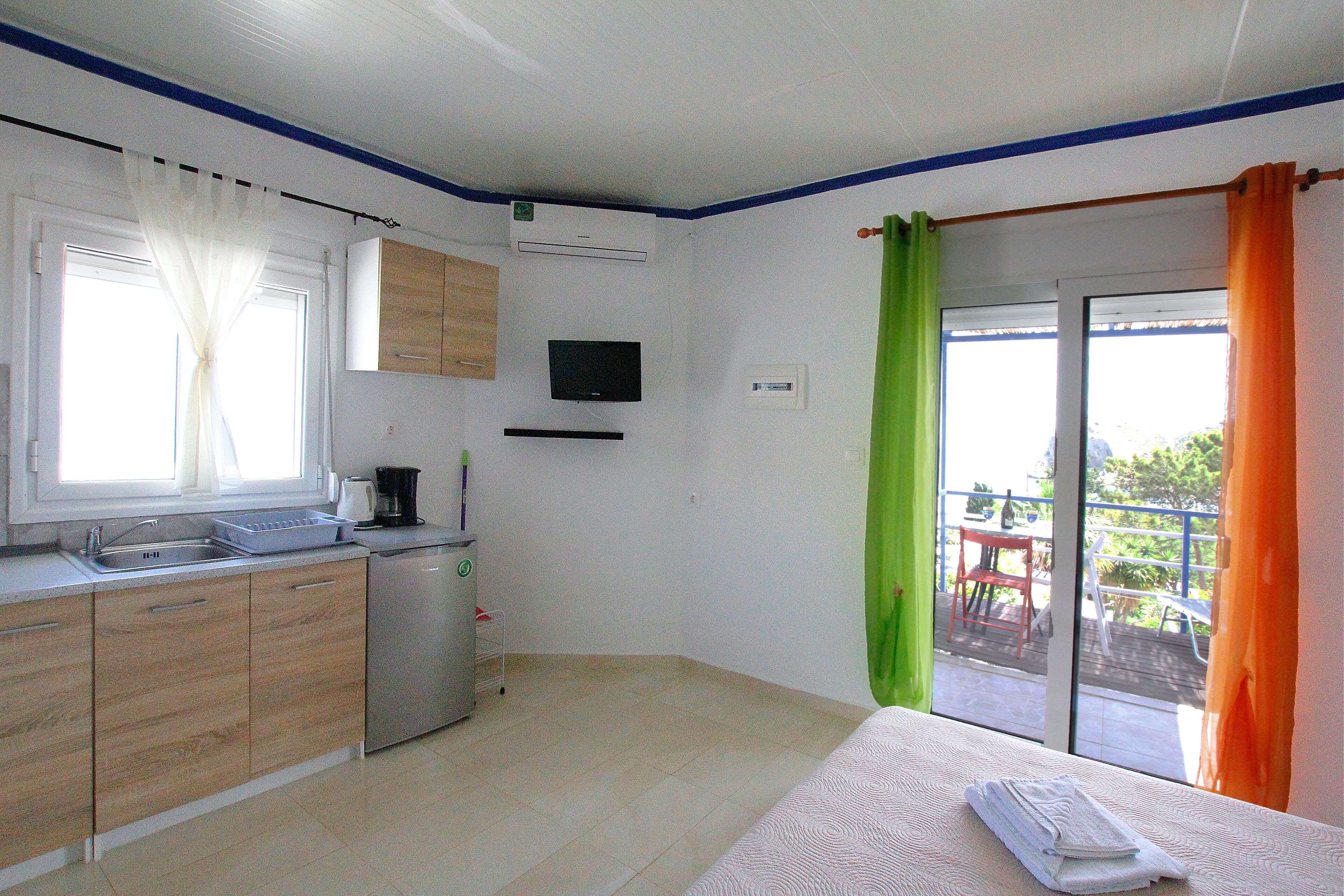 A compact hotel room with a kitchenette, a bed, a TV, and a balcony with a view.