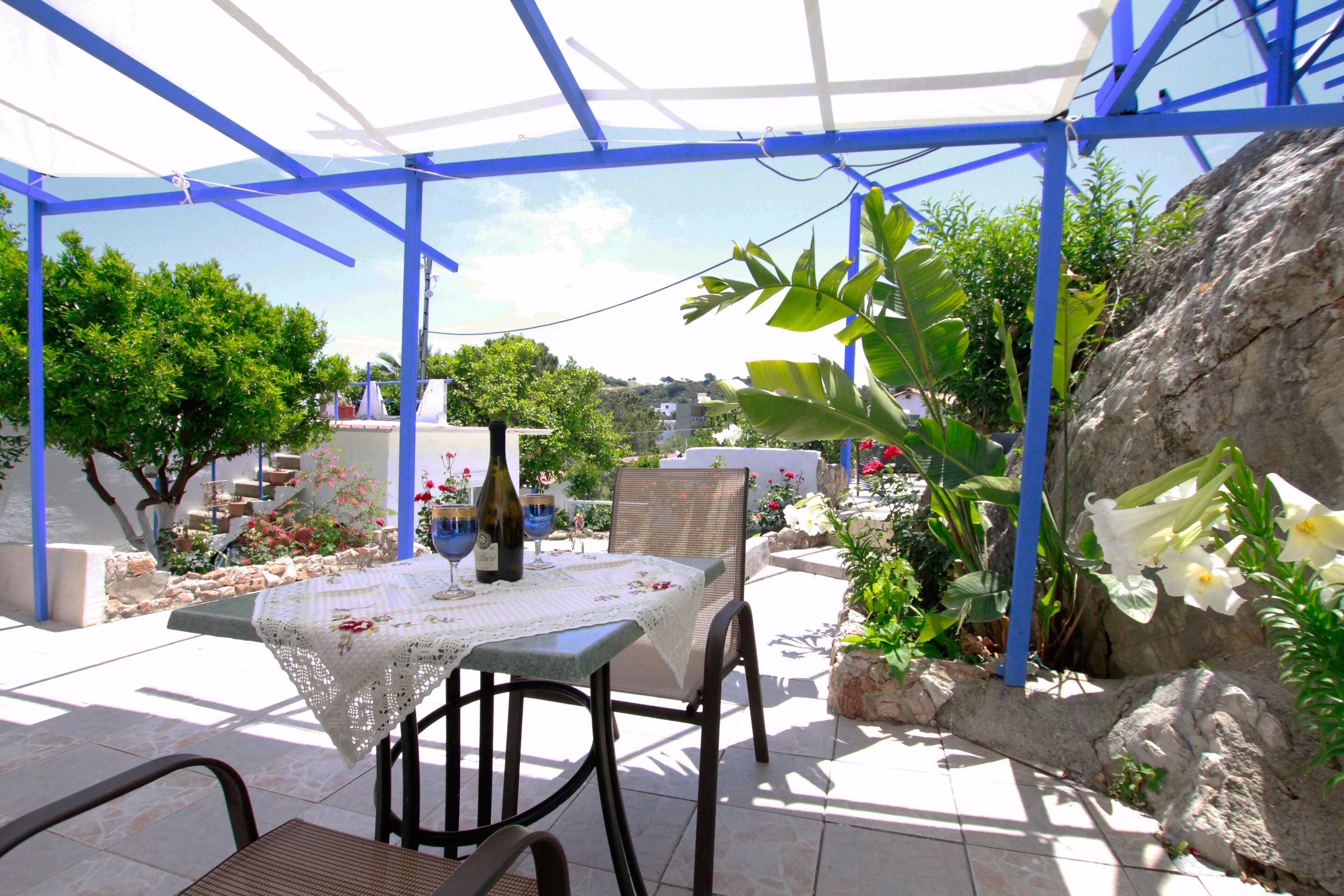 Un patio al aire libre con una mesa dispuesta para dos personas, rodeado de vegetación y un muro de piedra.