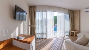 A hotel room with a TV mounted on the wall, a wooden desk with a remote control, a white sofa, and a view of a pool through sheer curtains. - Ponz Boutique Hotel (Kusadasi)