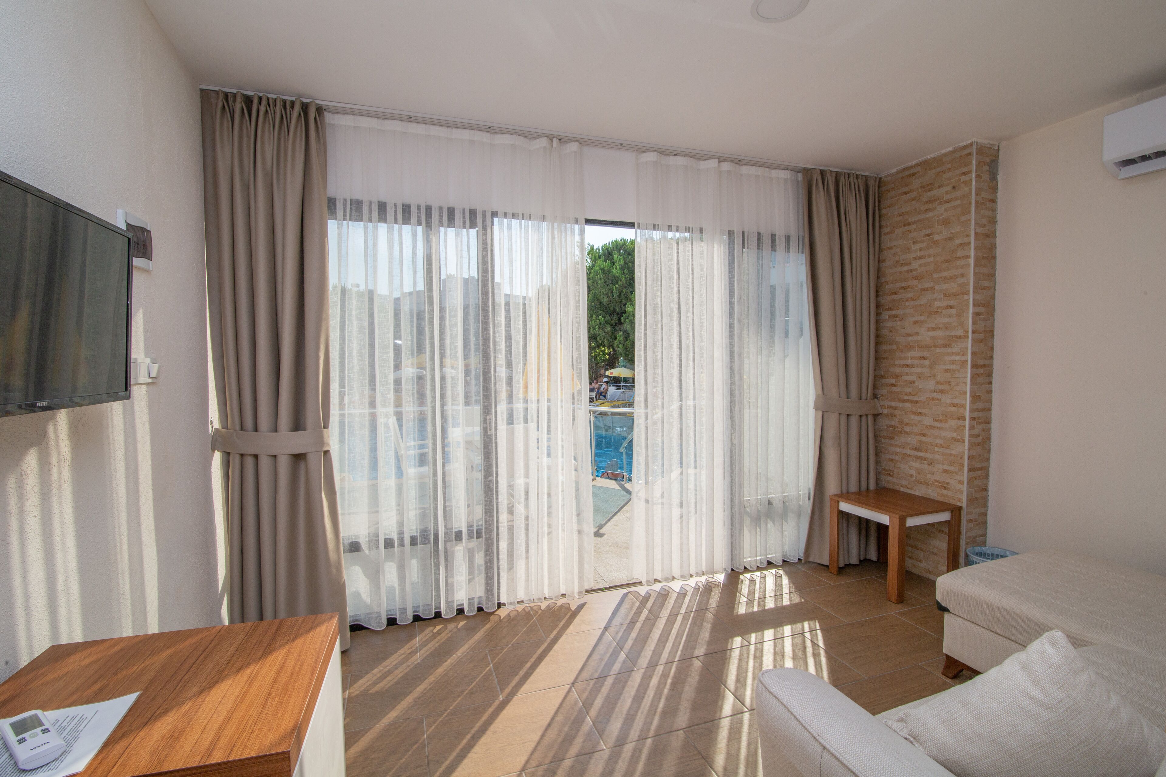 A hotel room with a large sliding glass door to a pool, a wooden table, a white sofa, and a TV mounted on the wall.