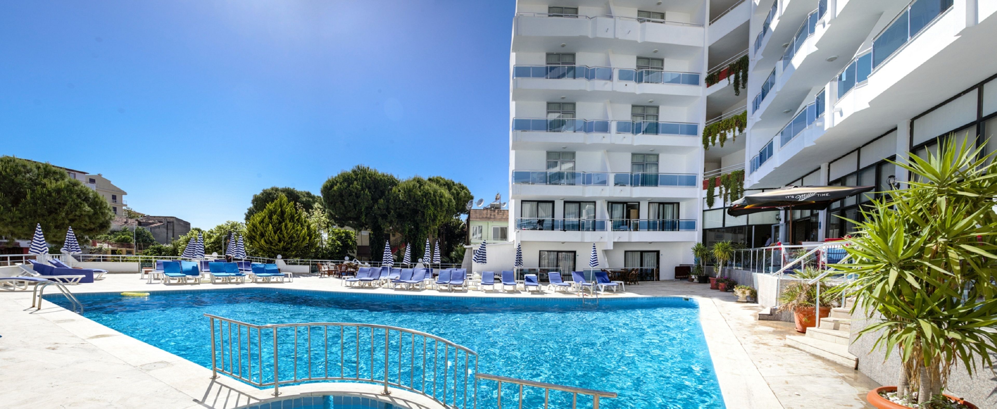 A swimming pool with lounge chairs and umbrellas, a multi-story building with balconies, and potted plants.