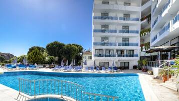 A swimming pool with lounge chairs and umbrellas, a multi-story building with balconies, and potted plants.