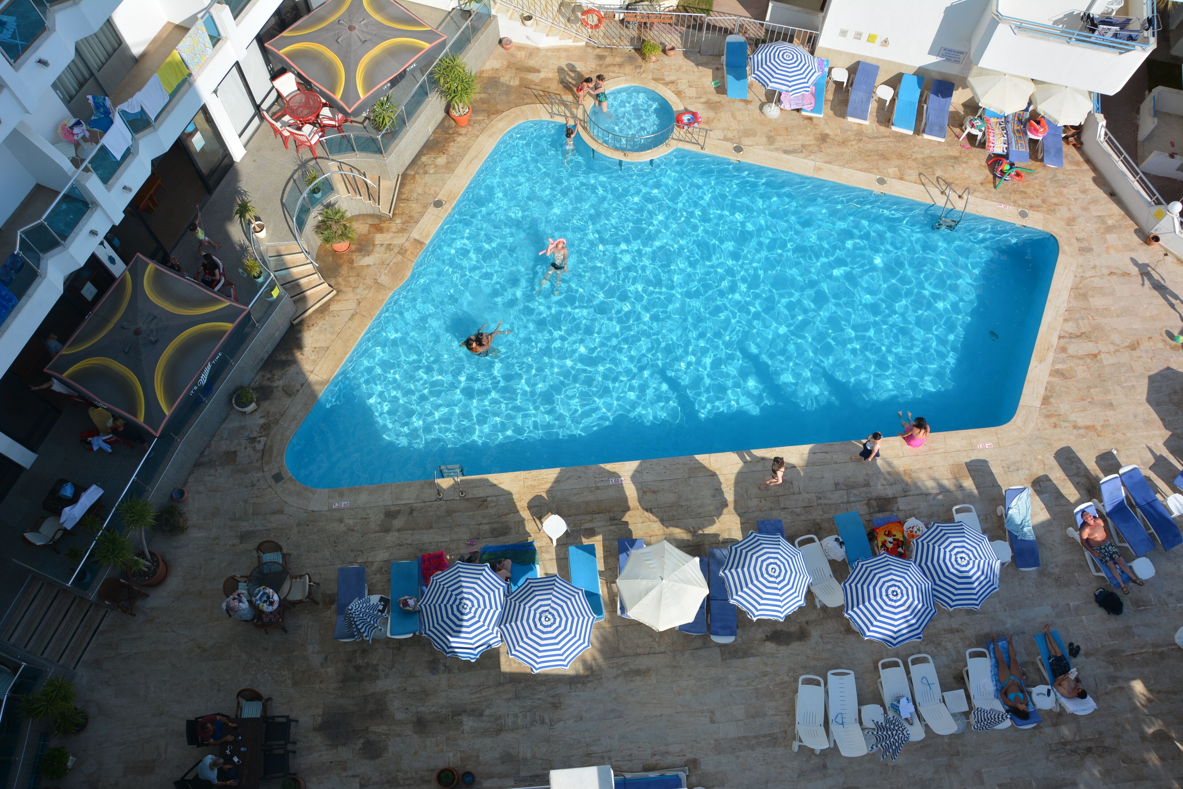 Ein Poolbereich auf dem Dach mit mehreren Liegestühlen und Sonnenschirmen, umgeben von einem Gebäude mit Balkonen und Pflanzen in Töpfen.