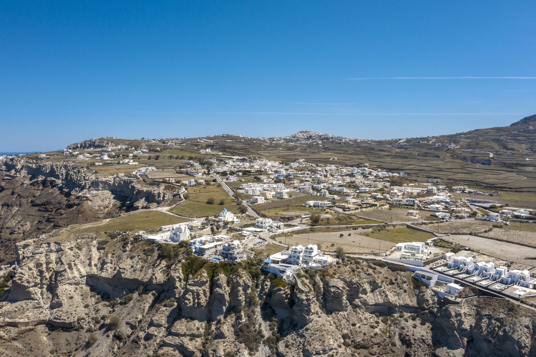 Vista aérea de un pueblo costero con edificios blancos, terreno rocoso y un cielo azul despejado.