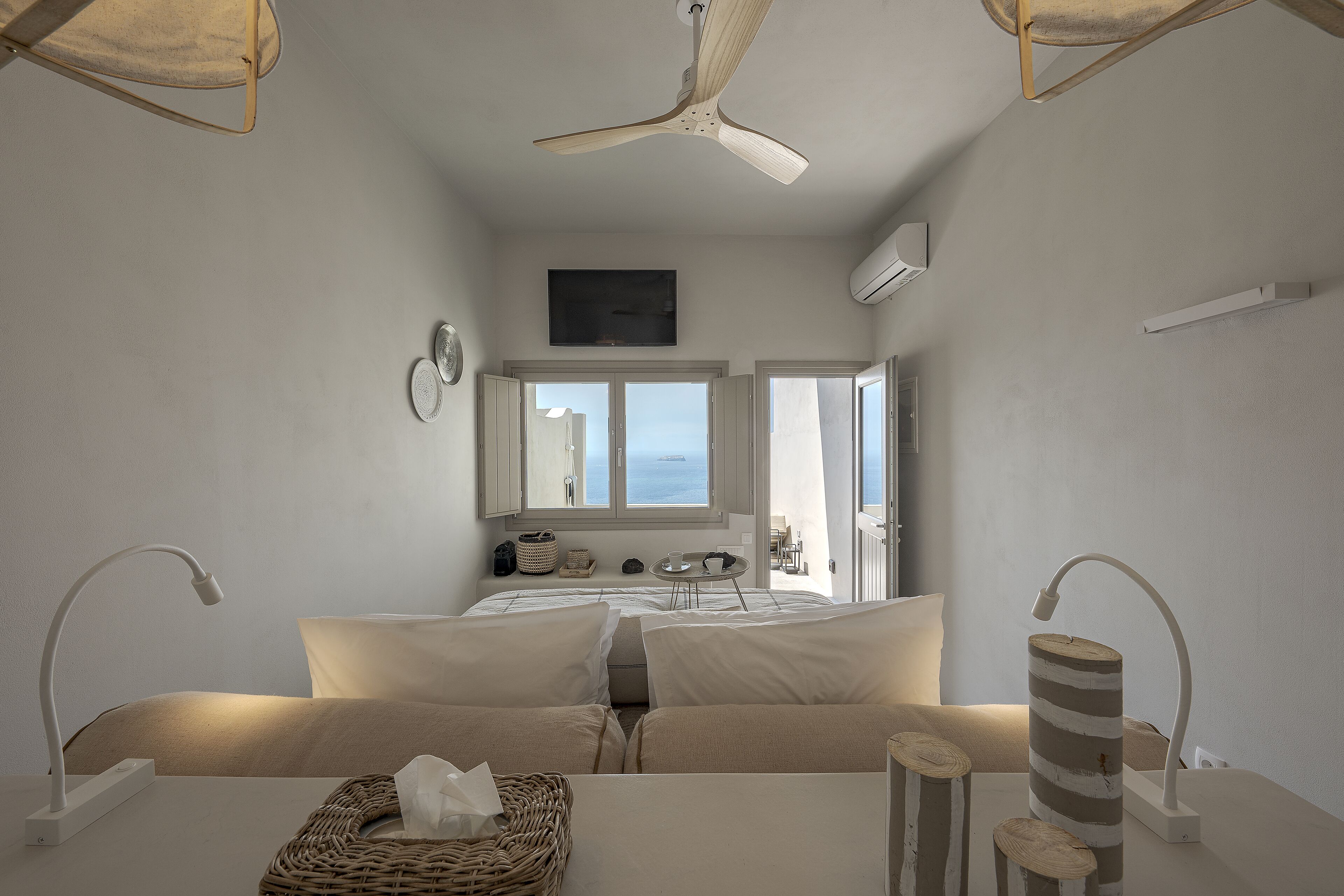 A bedroom with a bed, bedside lamps, a ceiling fan, a window, and a view of the sea.