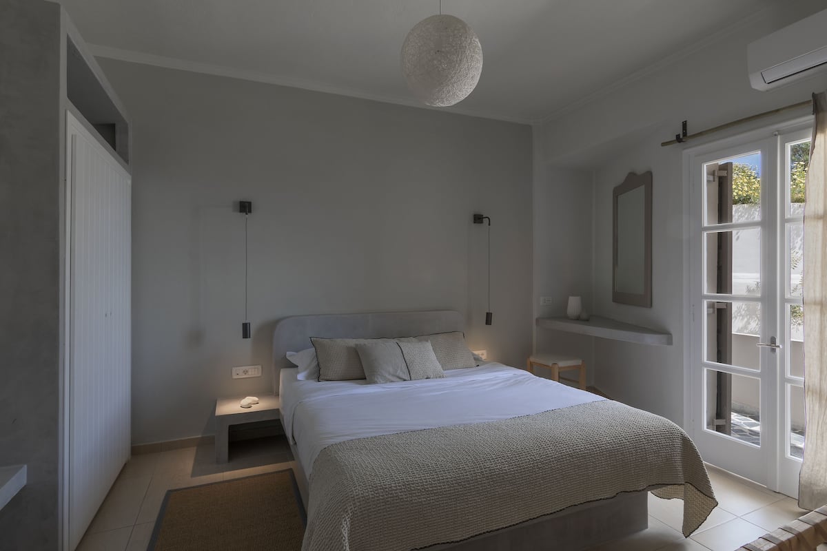 Un dormitorio con cama, mesitas de noche, lámpara colgante, ventana con cortinas y una puerta.
