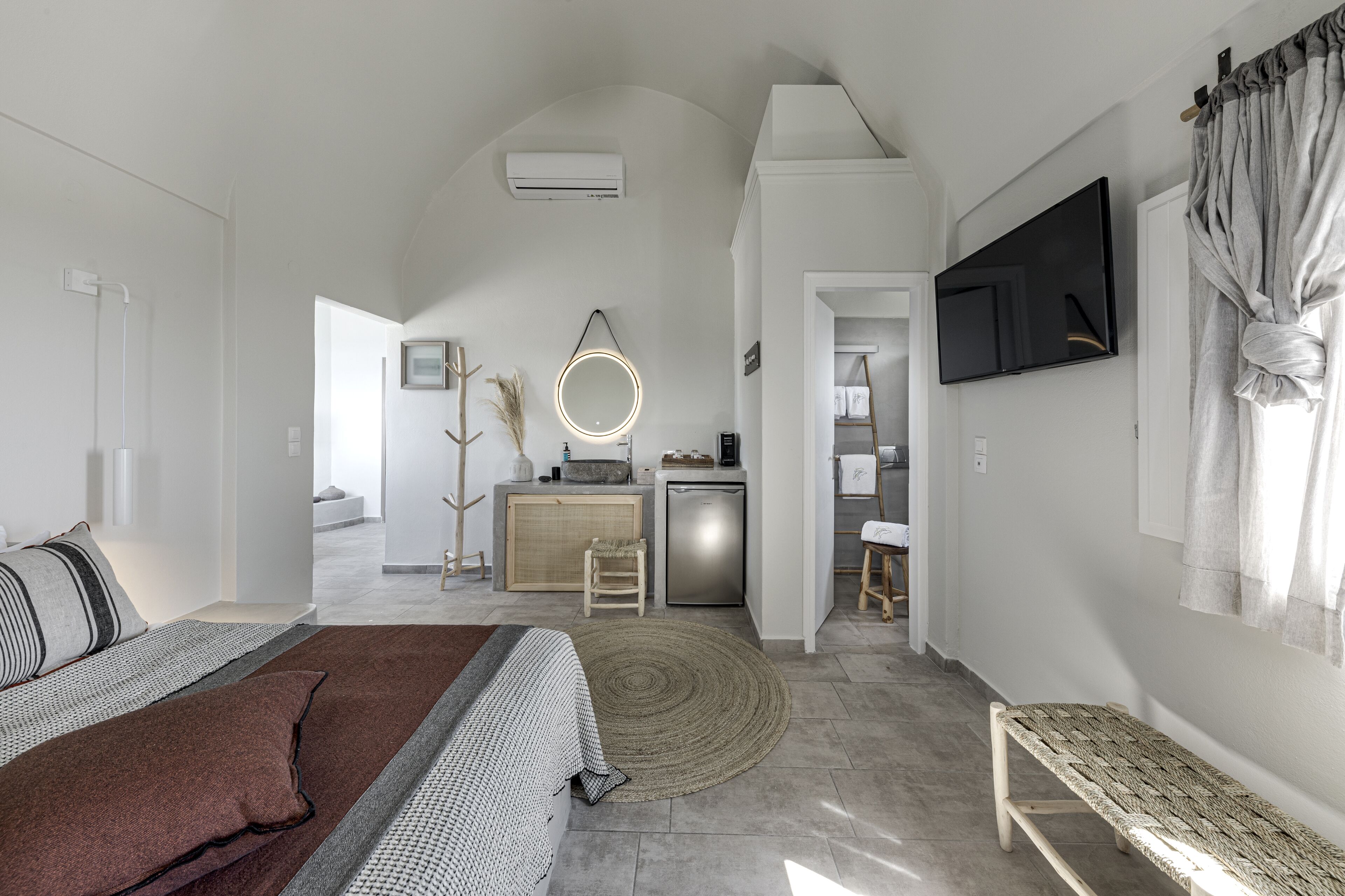 Un dormitorio moderno con cama, mesita de noche y un espejo grande. Se observa una kitchenette al fondo con un lavamanos y un refrigerador pequeño.