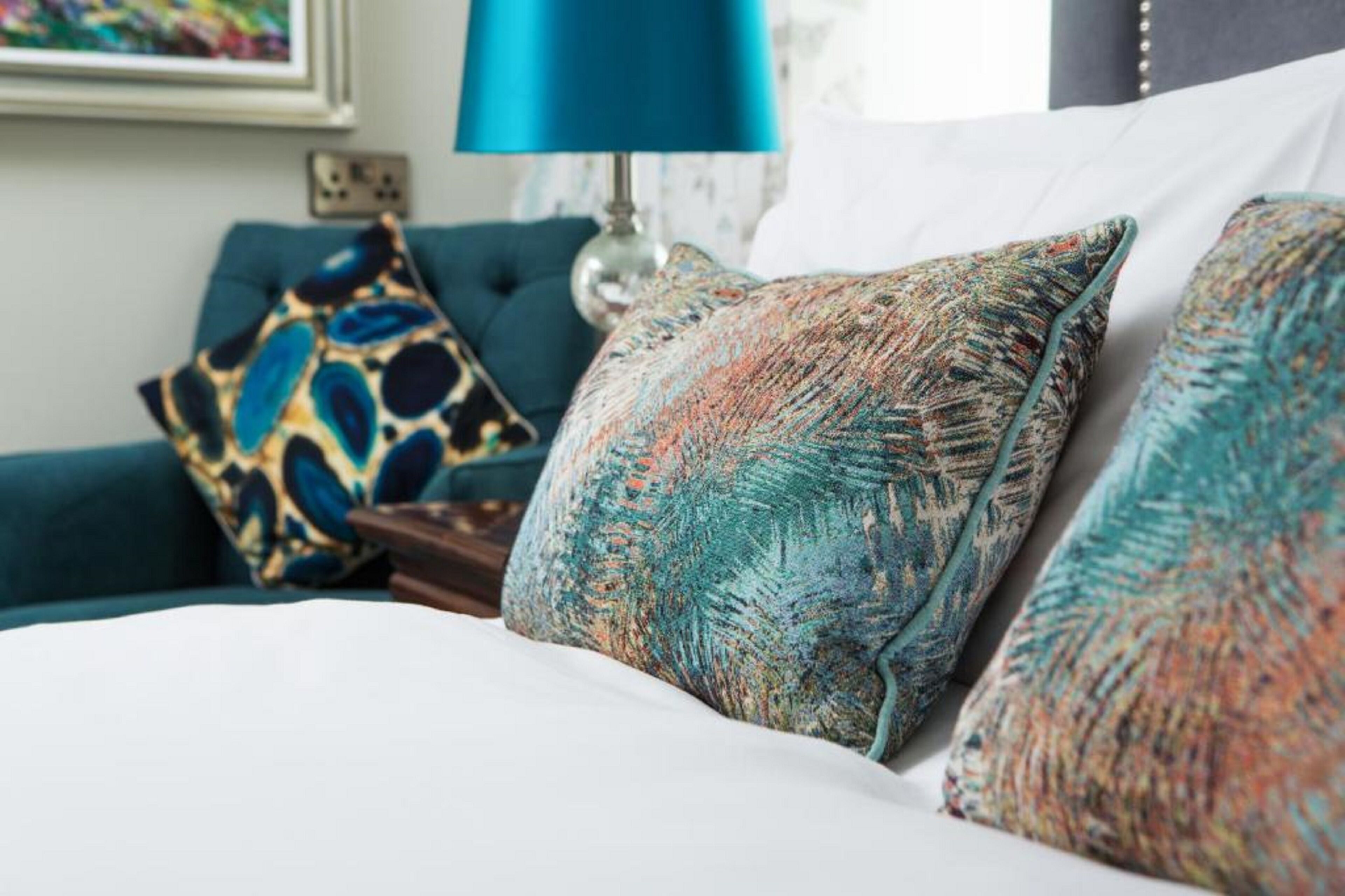 A bed with patterned pillows, a teal sofa, and a blue lamp in a hotel room.