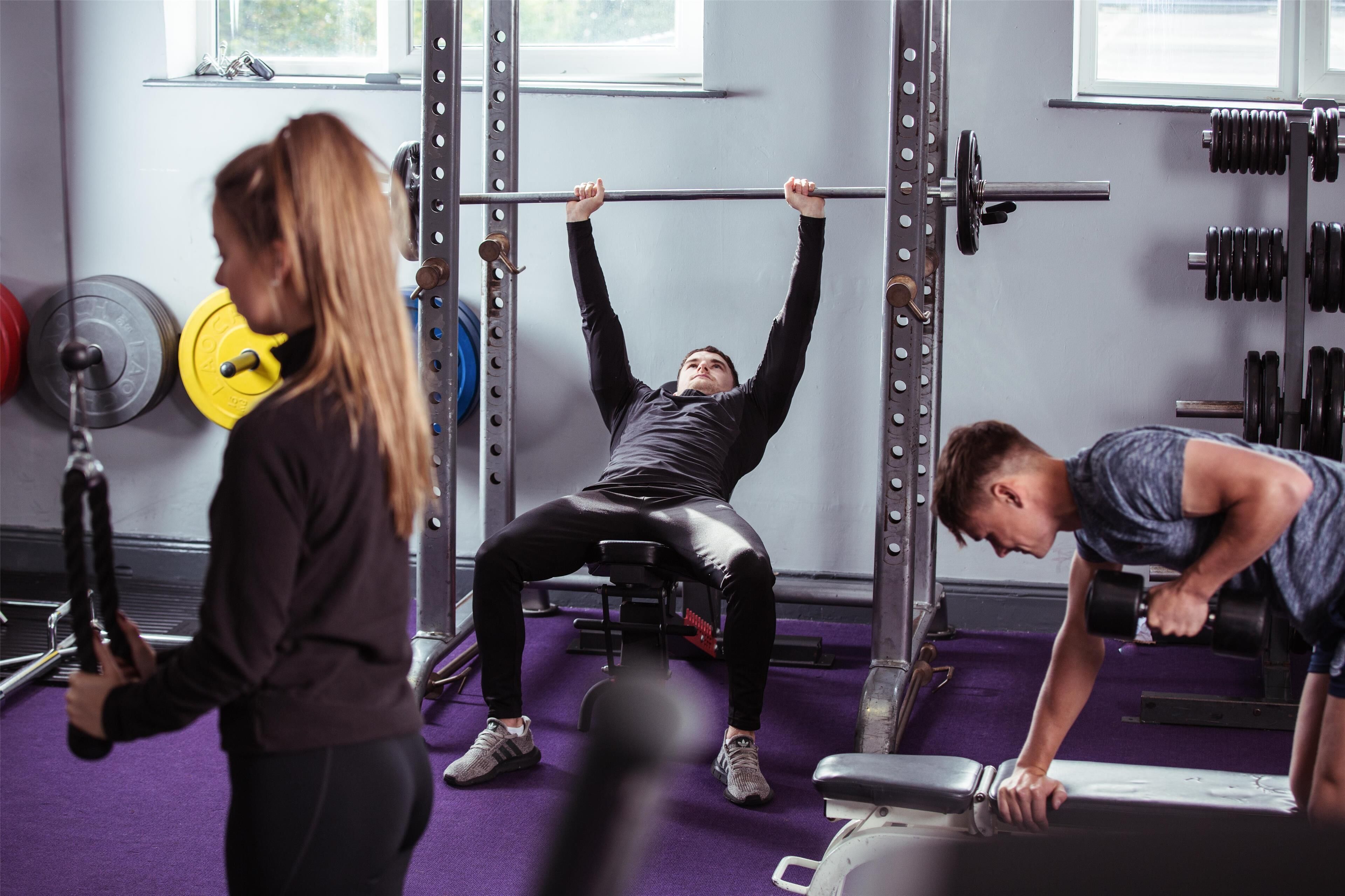 Un gimnasio con equipo de levantamiento de pesas, una persona haciendo press de banca y otra realizando extensiones de tríceps.