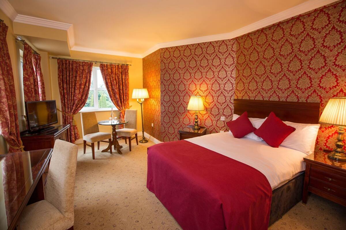 Quarto de hotel com uma cama grande, televisão, secretária com duas cadeiras e janela com cortinas.