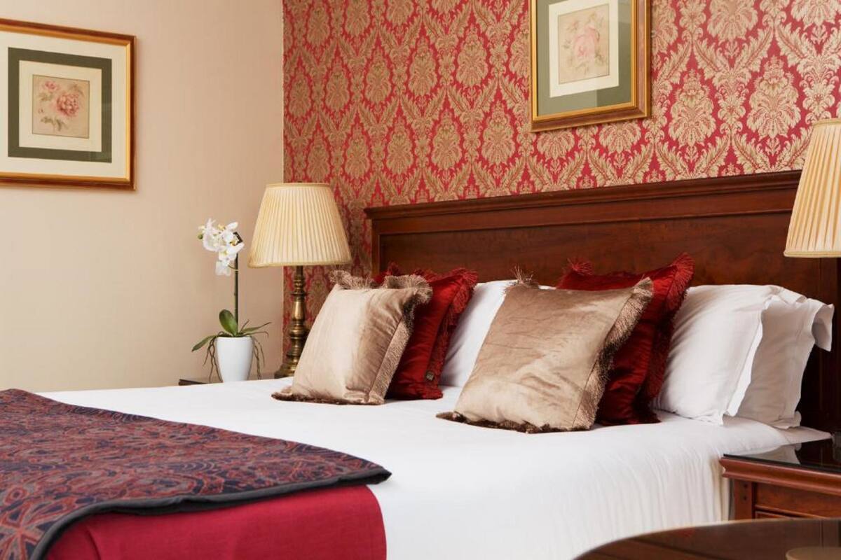 Uma cama bem arrumada com colcha de padrão vermelho e branco, cabeceira de madeira e duas luminárias de cabeceira.