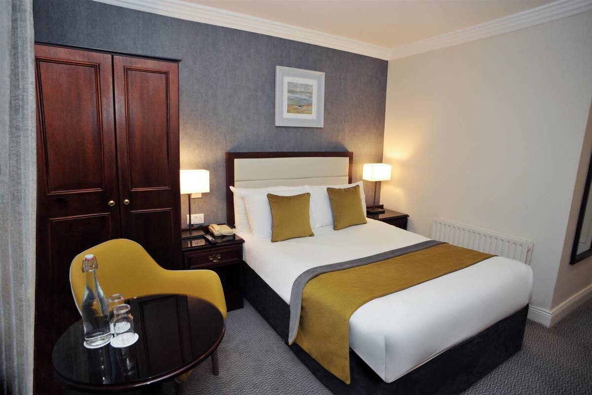 Quarto de hotel com uma cama grande, duas luminárias de cabeceira, uma pequena mesa com uma cadeira e um guarda-roupa de madeira espaçoso.