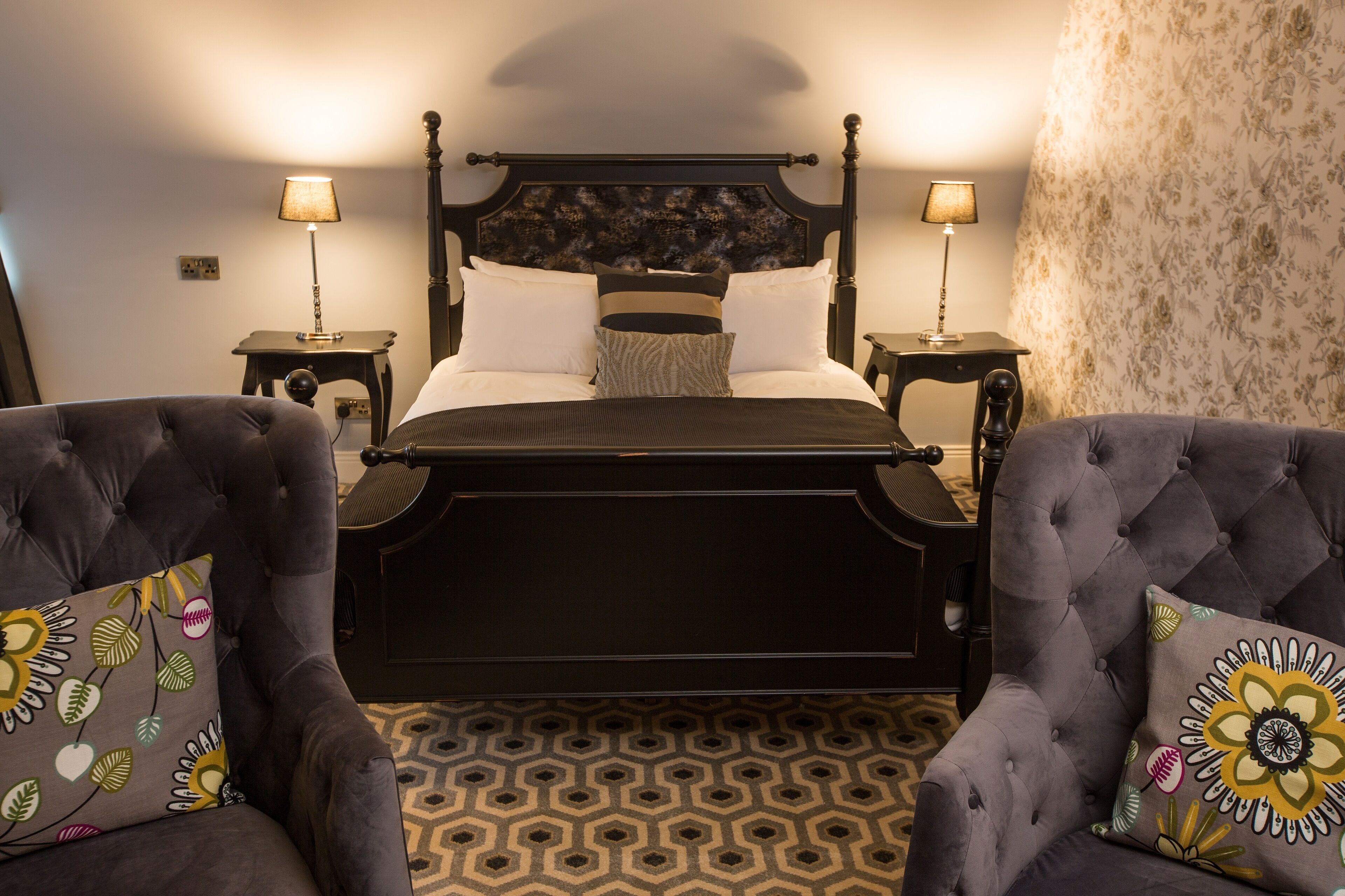 A neatly arranged bedroom with a four-poster bed, two bedside tables with lamps, and two tufted armchairs with floral-patterned cushions.