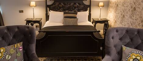 A neatly arranged bedroom with a four-poster bed, two bedside tables with lamps, and two tufted armchairs with floral-patterned cushions.