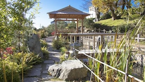 Un pabellón japonés tradicional con techo de tejas, rodeado por un sendero de piedra, plantas de bambú y un puente de piedra.